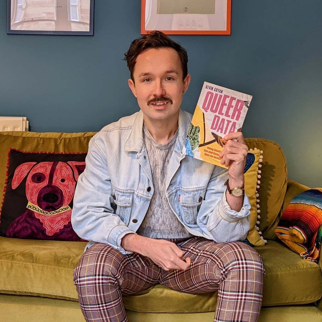 Kevin wearing a denim jacket and tartan trousers, holding a copy of Queer Data.